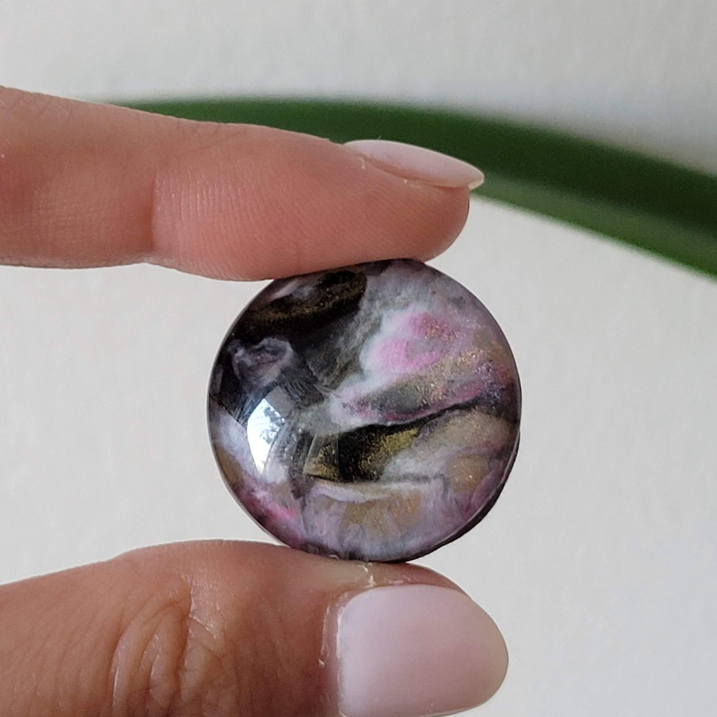 hand-painted circular glass magnet with pink, black, white and gold colors swirled, held in a hand