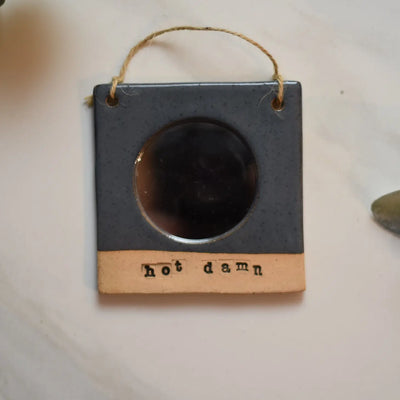 Ceramic frame with circular mirror and the words "hot damn" engraved at the bottom; hung on a twine string