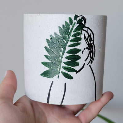 Concrete planter with hand painted silhouette of a woman from the side holding a leaf in green covering her face, held in a hand for reference, fits inside a palm