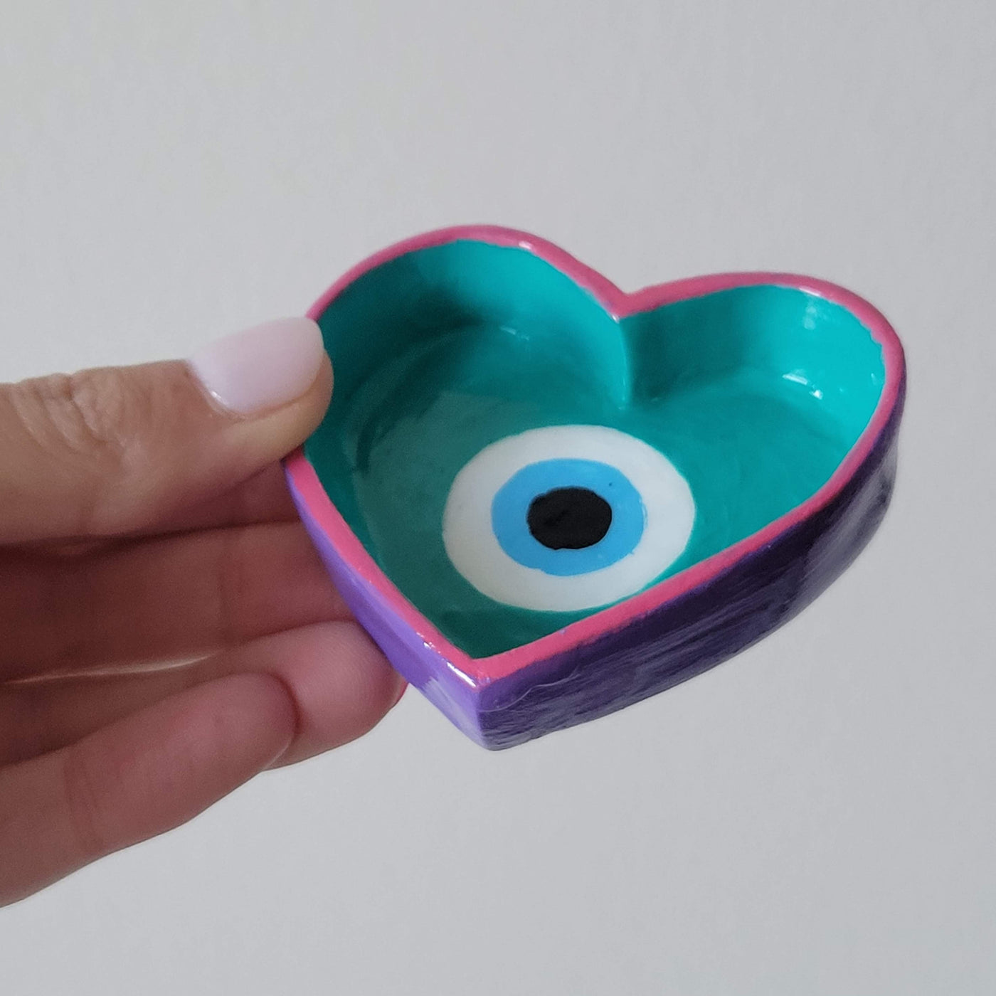 Evil eye painted in a heart shaped ring dish with pink, teal, blue, black and white, held in a hand to show the sides that are painted purple