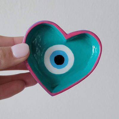 Evil eye painted in a heart shaped ring dish with pink, teal, blue, black and white, held in a hand