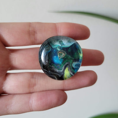 hand-painted circular glass magnet with shades of blue and green swirled, shown in a hand