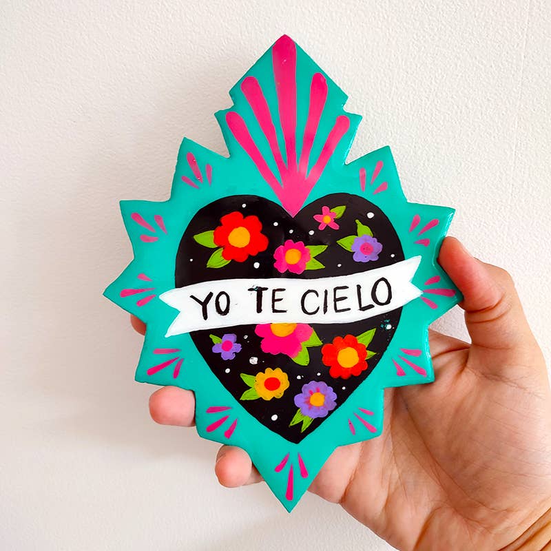 Wall hanging in the shape of a Mexican heart with "yo tel cielo" painted in caps in the center, held in the palm of a hand