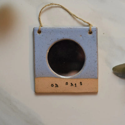 Ceramic frame with circular mirror and the words "oh shit" engraved at the bottom; hung on a twine string