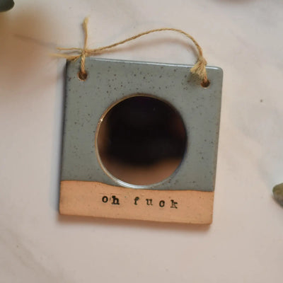Ceramic frame with circular mirror and the words "oh fuck" engraved at the bottom; hung on a twine string