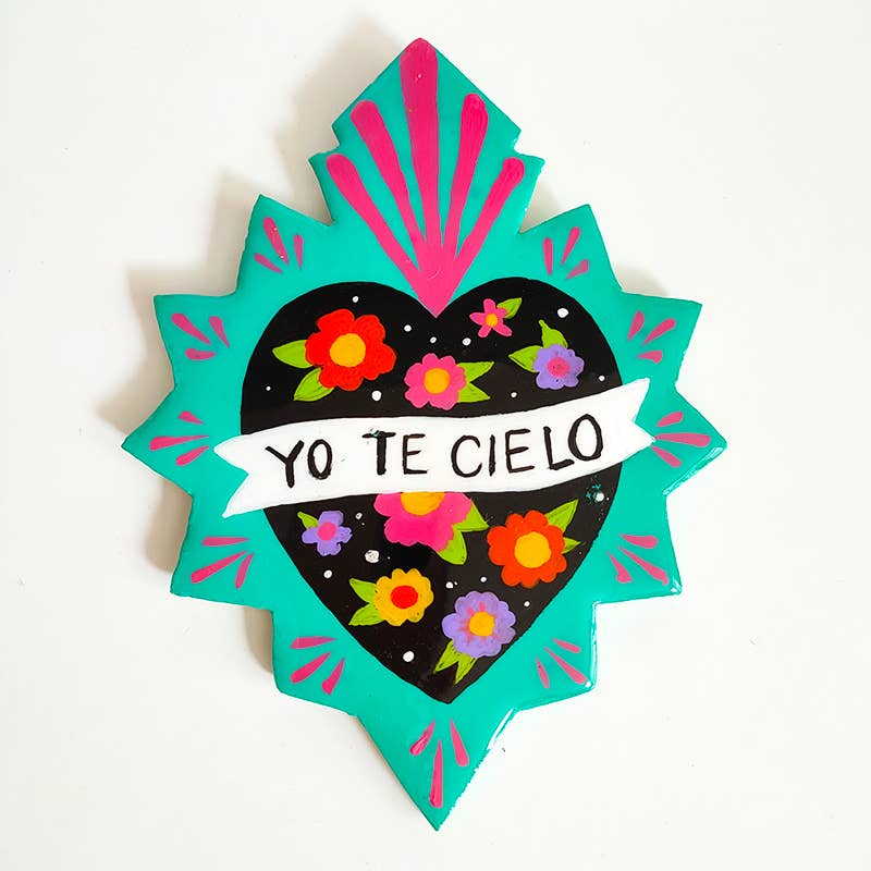 Wall hanging in the shape of a Mexican heart with "yo tel cielo" painted in caps in the center, colors are teal and pink with a black heart in the center and colorful flowers in the heart