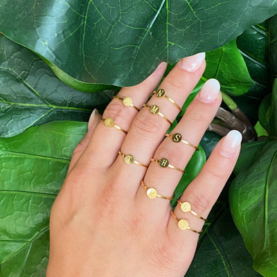 assortment of 18k gold plated stainless steel monogram rings on a model hand against leaves