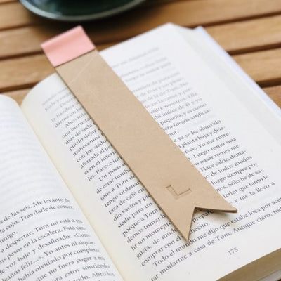 Tan leather bookmark with light pink color trim at top, and monogrammed "L" engraved towards bottom of the bookmark lying in an open book; bottom of bookmark is cut with two diagonals, forming the bottom of a tag or flag